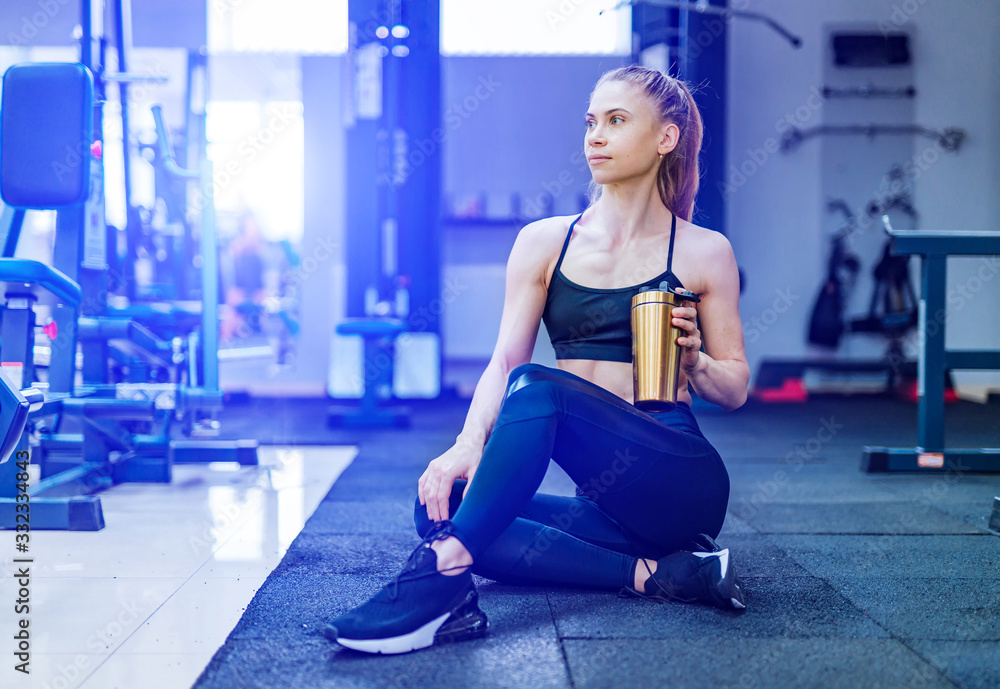 Fitness woman drinking water. Athletic girl sitting and resting on the floor in gym. Woman exercise workout in fitness gym breaking relax and holding protein shake bottle during training. Fitness woman drinking water. Athletic girl sitting and resting on the floor in gym. Woman exercise workout in fitness gym breaking relax and holding protein shake bottle during training.