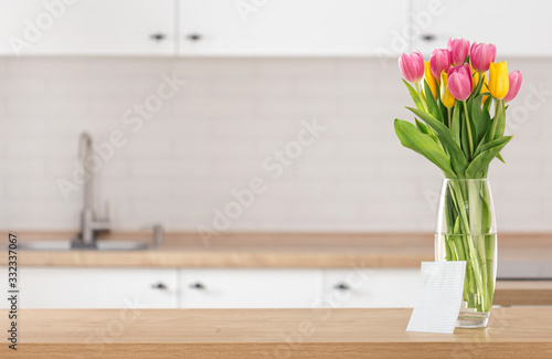 Bouquet of beautiful tulips on kitchen table with blank sheet of paper for your message on blur kitchen background with place for montage product display