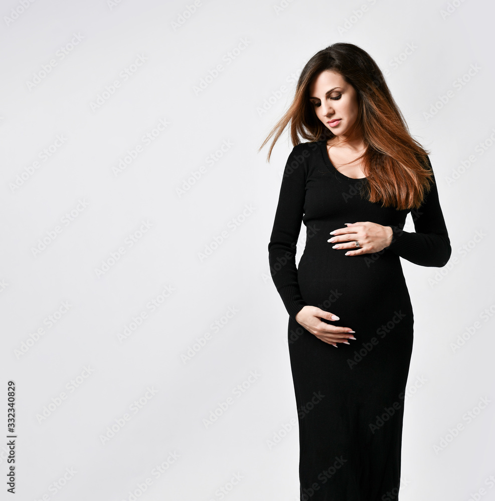 Brunette pregnant woman in black tight dress. She put hand on her belly while posing sideways isolated on white. Close up Brunette pregnant woman in black tight dress. She put hand on her belly while posing sideways isolated on white. Close up