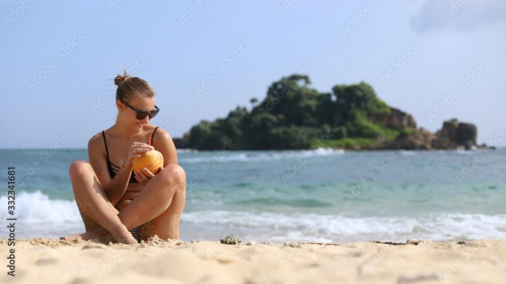 White young beautiful girl in a swimsuit and sunglasses drinks coconut water sitting on the sand at the beach. The coast of the ocean. Happiness. Smile. Relaxation. Tourism. White young beautiful girl in a swimsuit and sunglasses drinks coconut water sitting on the sand at the beach. The coast of the ocean. Happiness. Smile. Relaxation. Tourism.