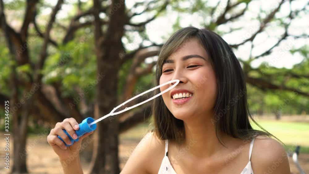South East Asian ethnic 20s woman blowing air bubbles in the park Young Happy girl wears stylish dress with trees in background. Outdoor summer weekend activity South East Asian ethnic 20s woman blowing air bubbles in the park Young Happy girl wears stylish dress with trees in background. Outdoor summer weekend activity