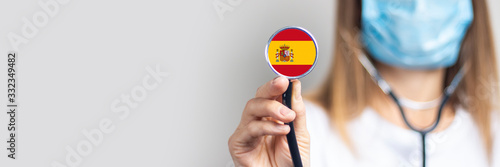 female doctor in a medical mask holds a stethoscope on a light background. Added flag of Spain. Concept medicine, level of medicine, virus, epidemic. Baner