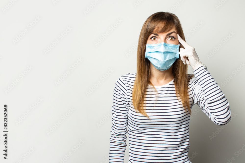 Young woman in a protective medical mask and latex gloves holds a finger at the temple on a light isolated background. Gesture think, be careful. Quarantine, remedies, coronavirus concept Young woman in a protective medical mask and latex gloves holds a finger at the temple on a light isolated background. Gesture think, be careful. Quarantine, remedies, coronavirus concept