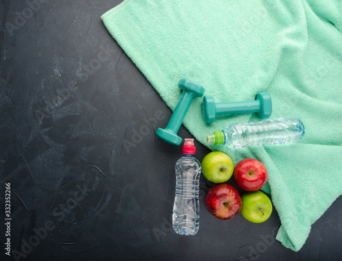 Healthy lifestyle concept. colored Apples dumbbells sport water bottles and turquoise towel on black concrete background