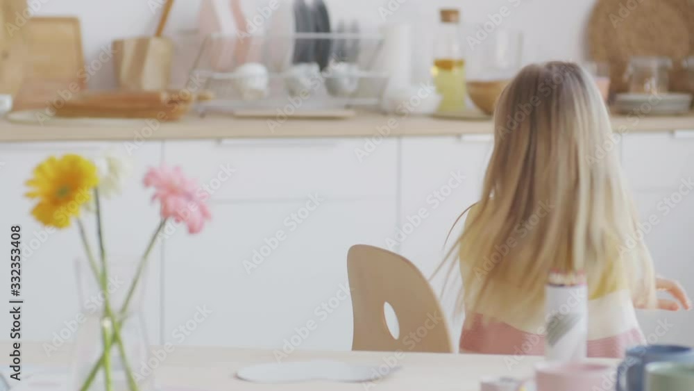 Medium shot of Caucasian cute girl with long blond hair sitting at kitchen table and spinning on chair Medium shot of Caucasian cute girl with long blond hair sitting at kitchen table and spinning on chair