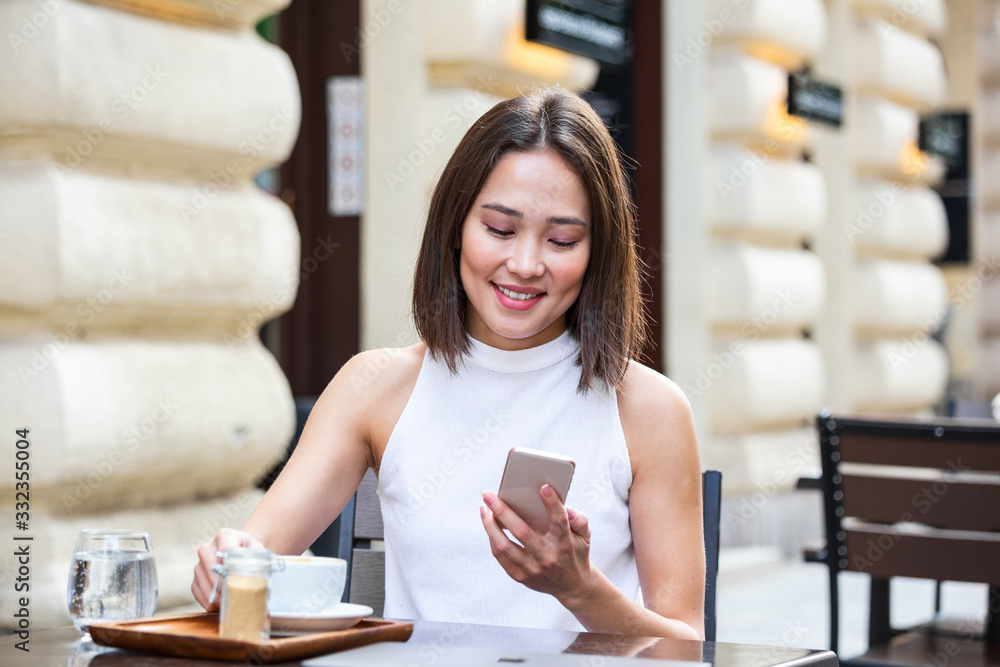 Smiling Asian woman drinking coffee and using her mobile phone. Satisfied female enjoying cup of coffee. Close up portrait of beautiful girl drinking coffee from a white mug in the coffee shop Smiling Asian woman drinking coffee and using her mobile phone. Satisfied female enjoying cup of coffee. Close up portrait of beautiful girl drinking coffee from a white mug in the coffee shop