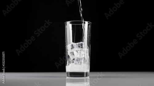 Pure fresh mineral water is poured into a transparent glass with ice cubes on a black background. Concept of stay at home and eat healthy food with soda and ice. Wide shot. Slow motion