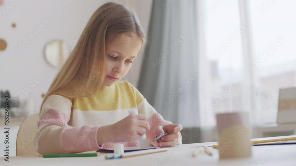 Medium shot of pretty Caucasian girl sitting at table and making greeting card for Mothers day using glue stick Medium shot of pretty Caucasian girl sitting at table and making greeting card for Mothers day using glue stick