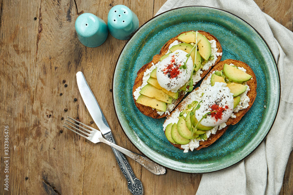 Trending food - avocado toast with ricotta, poached egg on rye bread in a ceramic plate on a wooden background. Healthy breakfast meal