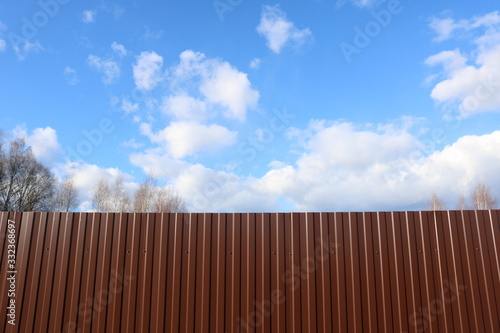 A texture of brown iron fence with tree tops behind it and a blue sky with white clouds. 