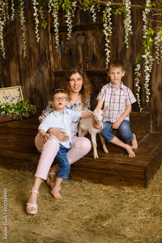 sitting young mother and two her sons kids with little white goat on dark wooden background with flowers and hay