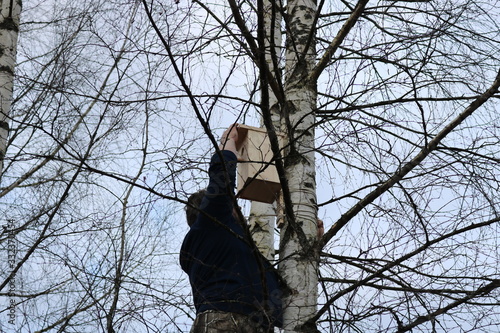 Bare branches of a birch tree through which you can see part of the figure of a man attaching the roof to a birdhouse hanging on this birch tree.