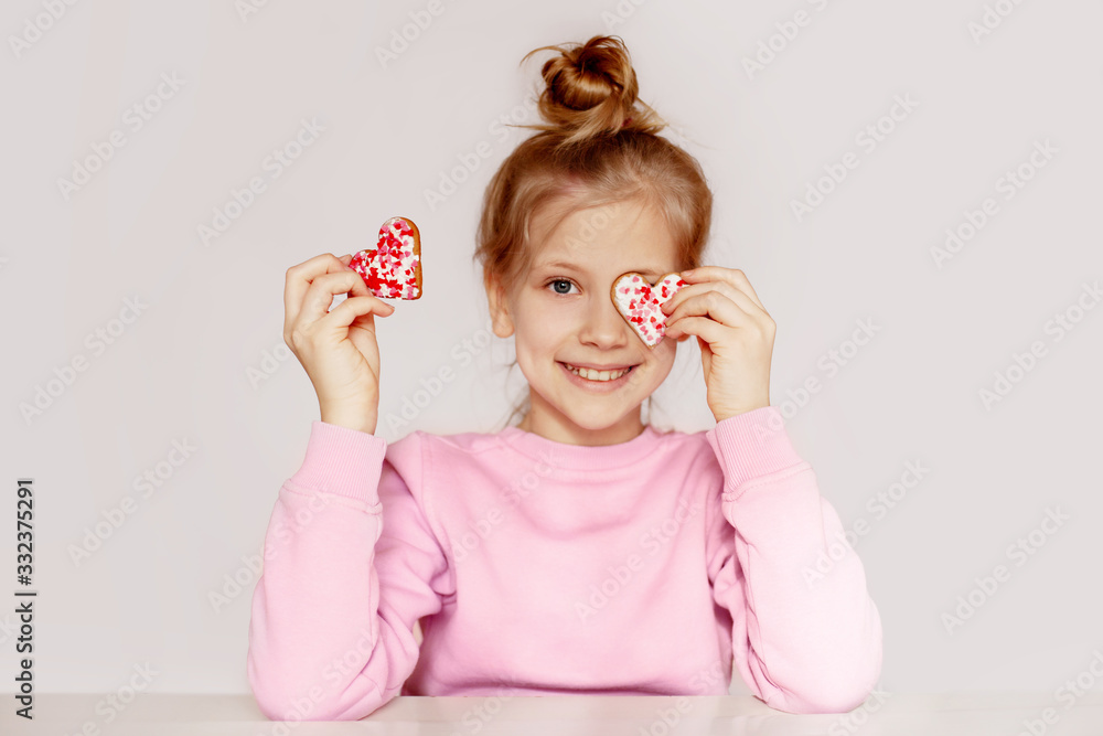 A girl in a pink sweater is holding gingerbread in the shape of a heart. The child closes his eyes with cookies