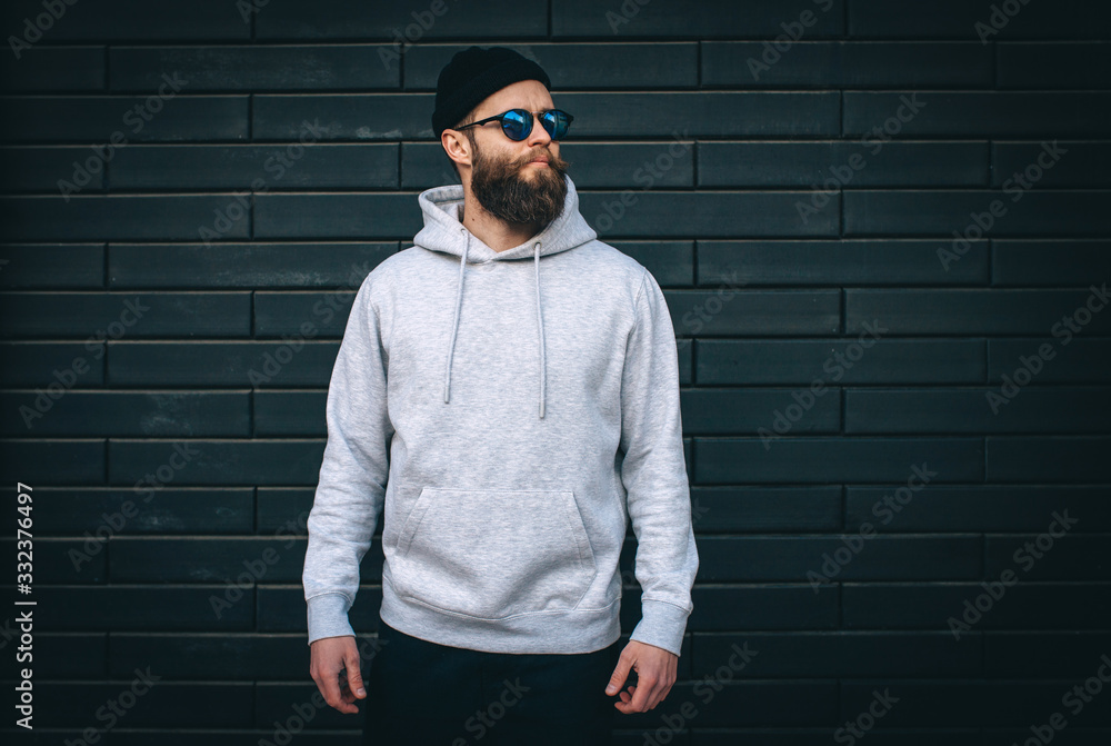 City portrait of handsome hipster guy with beard wearing gray blank hoodie or hoody and hat with space for your logo or design. Mockup for print City portrait of handsome hipster guy with beard wearing gray blank hoodie or hoody and hat with space for your logo or design. Mockup for print