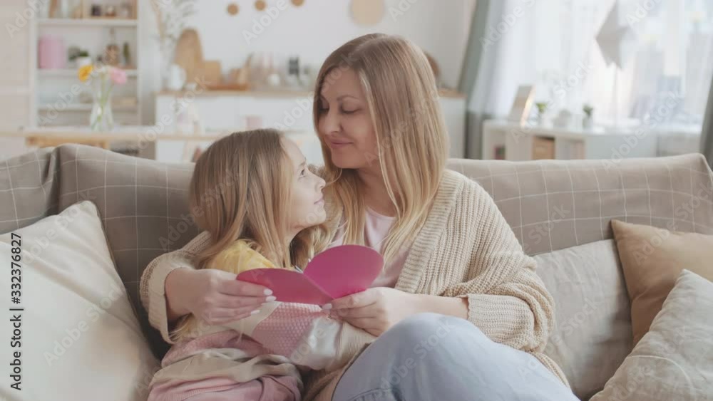Medium shot of mother greeting her little daughter for handmade heart-shaped card by hugging and kissing her Medium shot of mother greeting her little daughter for handmade heart-shaped card by hugging and kissing her