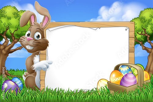 Easter bunny rabbit peeking around a sign and pointing with Easter eggs and basket background cartoon
