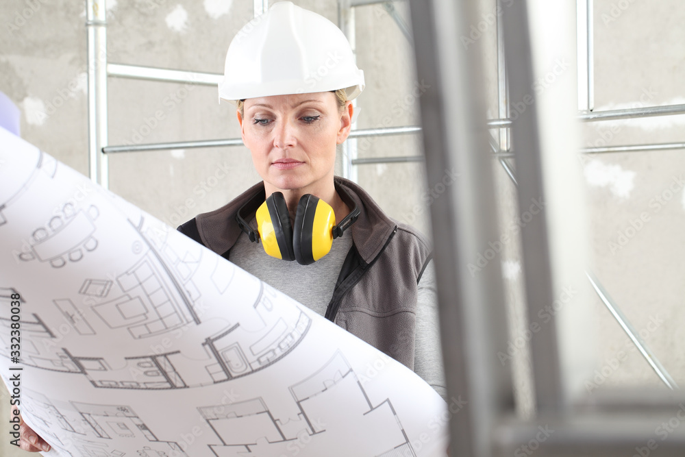 woman construction worker builder looking at bluprint, wearing helmet and hearing protection headphones in building site indoors background woman construction worker builder looking at bluprint, wearing helmet and hearing protection headphones in building site indoors background