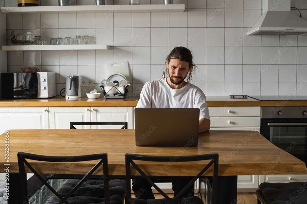 Freelancer working from home on the kitchen and using laptop. Bearded man working with a laptop and reading bad news. Handsome successful self entrepreneur sitting and working at his modern home. Freelancer working from home on the kitchen and using laptop. Bearded man working with a laptop and reading bad news. Handsome successful self entrepreneur sitting and working at his modern home.