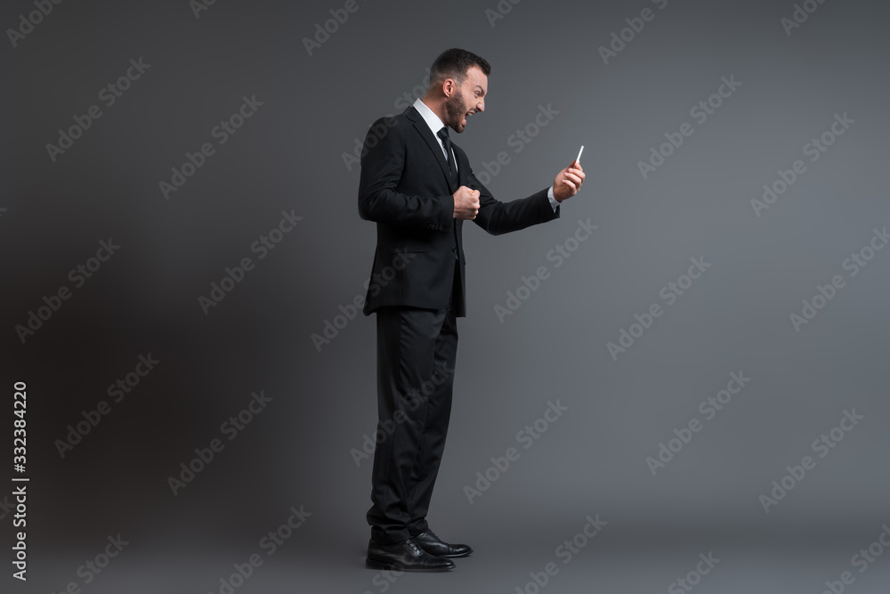profile of angry businessman in suit screaming while looking at smartphone on grey