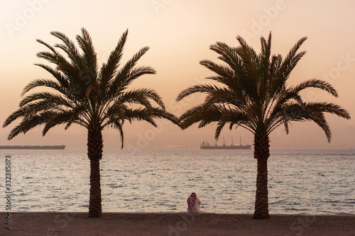 Arabian man sitting on the coast between two palms. Sunset. Aqaba  Jordan.