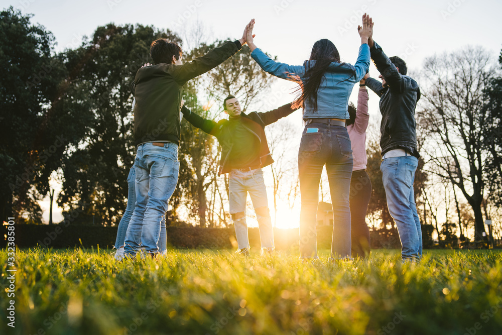 Fototapeta premium Group of young friends at park at sunset in circle with hands upwards - Teens in a moment of unity, fraternity, strength and team building