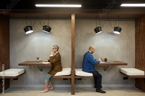 Side view full length portrait of two adult people separated by wall while sitting in separate cafe booths, copy space
