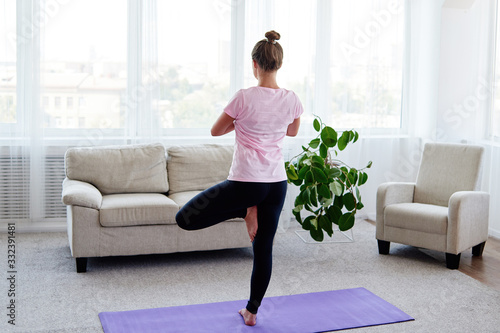 Fototapeta Naklejka Na Ścianę i Meble -  Portrait of young woman practicing balance yoga asana Vrikshasana at home indoor, copy space, back view. Girl doing tree pose, full length. Relaxing and doing yoga. Wellness and healthy lifestyle