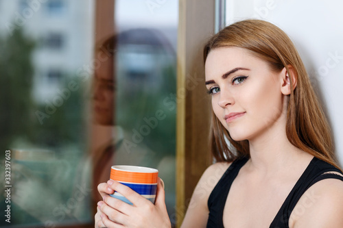 Young woman sitting with cup at windowsill at home in rainy day.