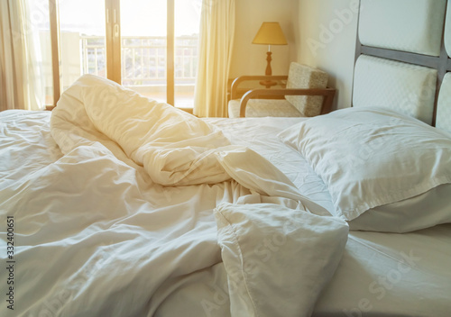 Messy white bedding sheet and white pillow after waking up in the morning with sunshine shining through window with curtains in to the bed room, waking up concept