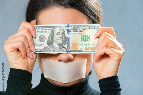 girl with mouth taped in white tape close-up portrait, holds a hundred-dollar bill in front of her face, closing her eyes. because of money he doesn’t see anything. selfishness and preemptiveness. USD photo