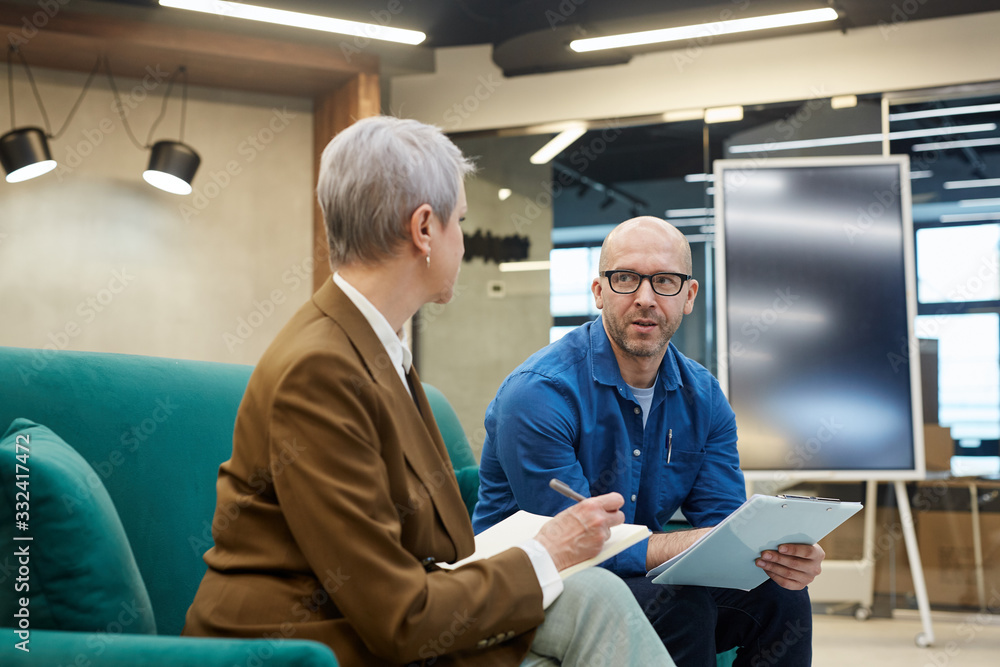Obraz premium Portrait of bald mature man discussing work project with female manager while sitting on couch in modern office interior, copy space