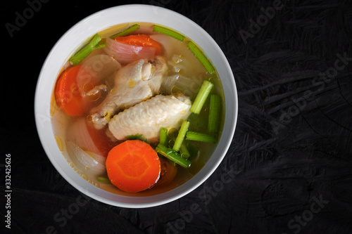 Bowl of chicken soup and chicken wings with carrots, onion and tomatoes on wood table. 