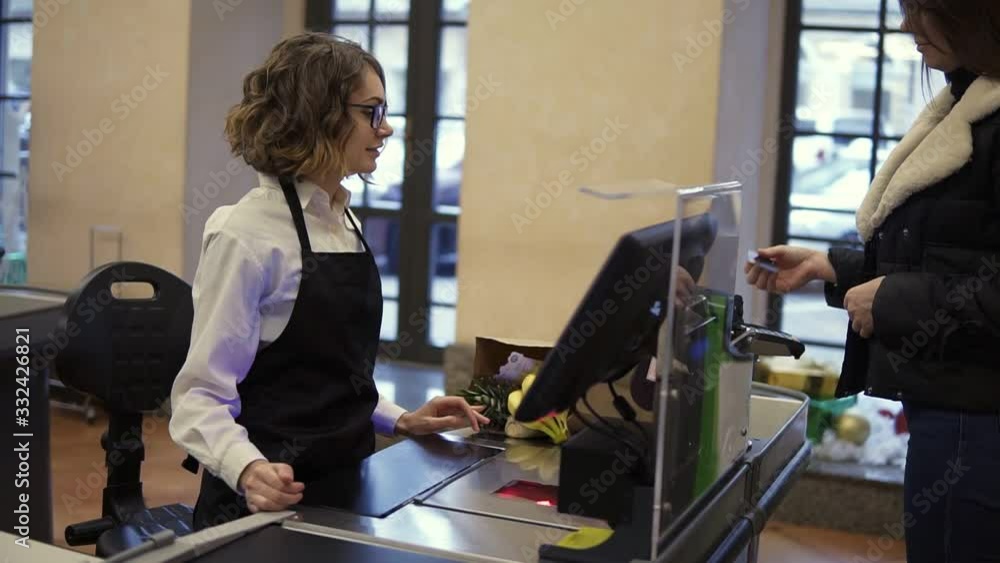 Smiling woman customer paying with her EC card at supermarket checkout and taking her brown paper bag with products from the counter. Female cashier standing in black apron