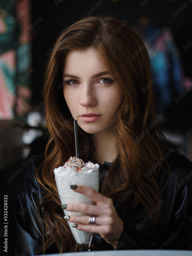 Close up shot of beautiful cunning redhead woman in black leather shirt sitting in trendy cozy city cafe near big window holding milkshake glass