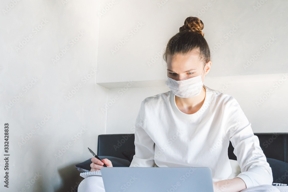 young woman in glasses working from home on laptop in bedroom, wearing face protective mask. quarantine and isolation because of coronavirus flu virus. young woman in glasses working from home on laptop in bedroom, wearing face protective mask. quarantine and isolation because of coronavirus flu virus.