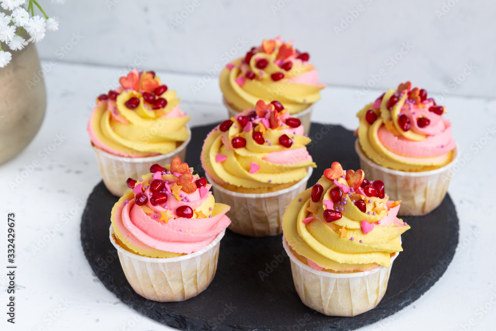 Traditional lemon muffins on a black tray and white background. With pink-yellow butter cream and pomegranate seeds.