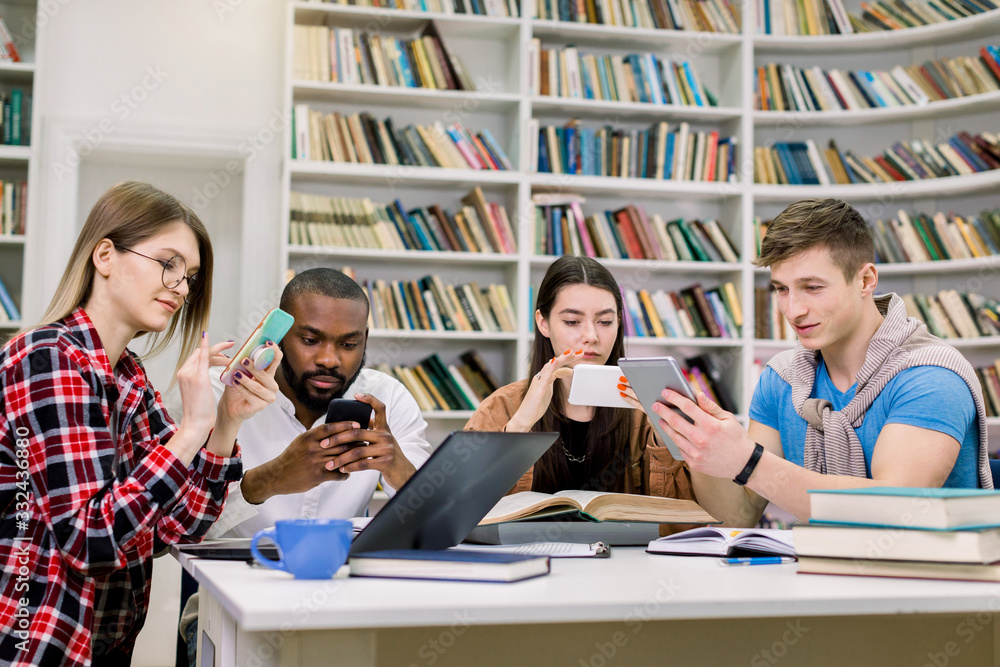 Four young friends students, boys and girls of mixed race, which are preparing for exams and searching needed information on their smartphones, sitting in the reading room in library Four young friends students, boys and girls of mixed race, which are preparing for exams and searching needed information on their smartphones, sitting in the reading room in library