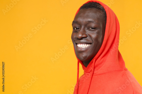 Side view of smiling young african american man guy in red streetwear hoodie with hood on head posing isolated on yellow wall background. People lifestyle concept. Mock up copy space. Looking camera. © ViDi Studio