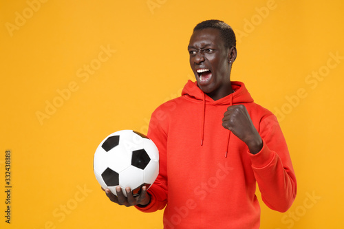 Screaming young african american man guy football player in red streetwear hoodie isolated on yellow wall background. Sport leisure lifestyle concept. Playing football hold soccer ball clenching fist. © ViDi Studio