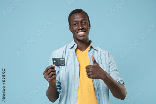 Smiling young african american man guy in casual shirt, yellow t-shirt posing isolated on blue wall background. People lifestyle concept. Mock up copy space. Hold credit bank card showing thumb up.