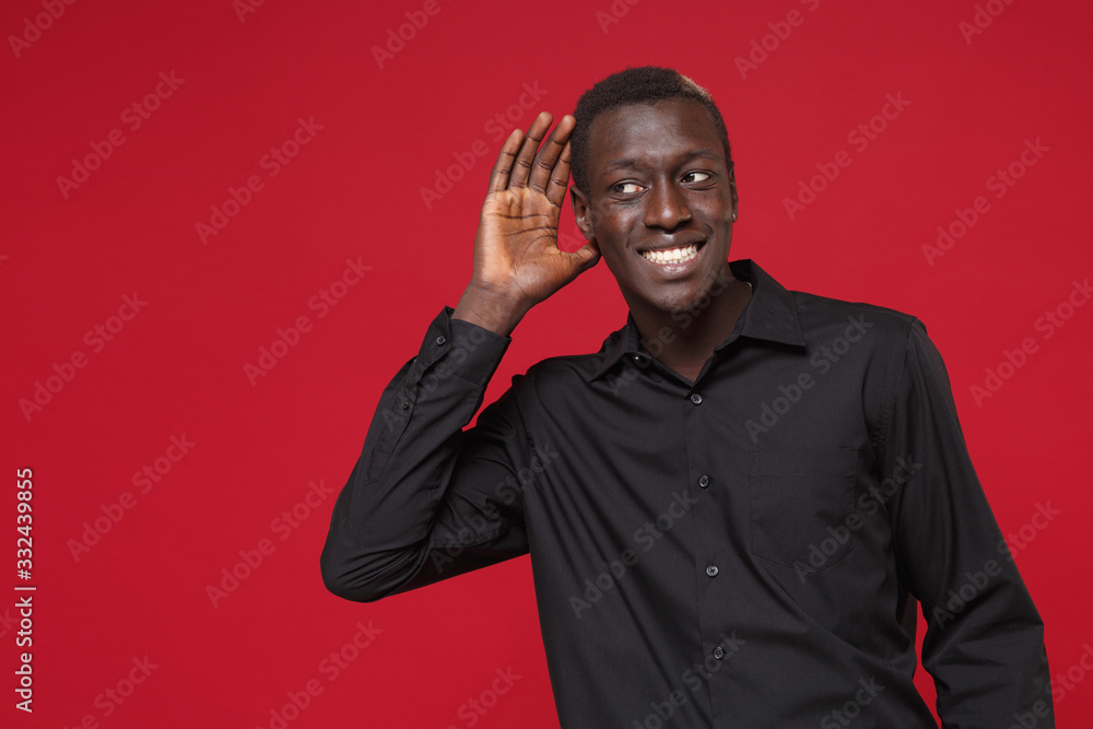 Smiling young african american man in classic black shirt posing isolated on red background studio portrait. People lifestyle concept. Mock up copy space. Try to hear you with hand gesture near ear. Smiling young african american man in classic black shirt posing isolated on red background studio portrait. People lifestyle concept. Mock up copy space. Try to hear you with hand gesture near ear.