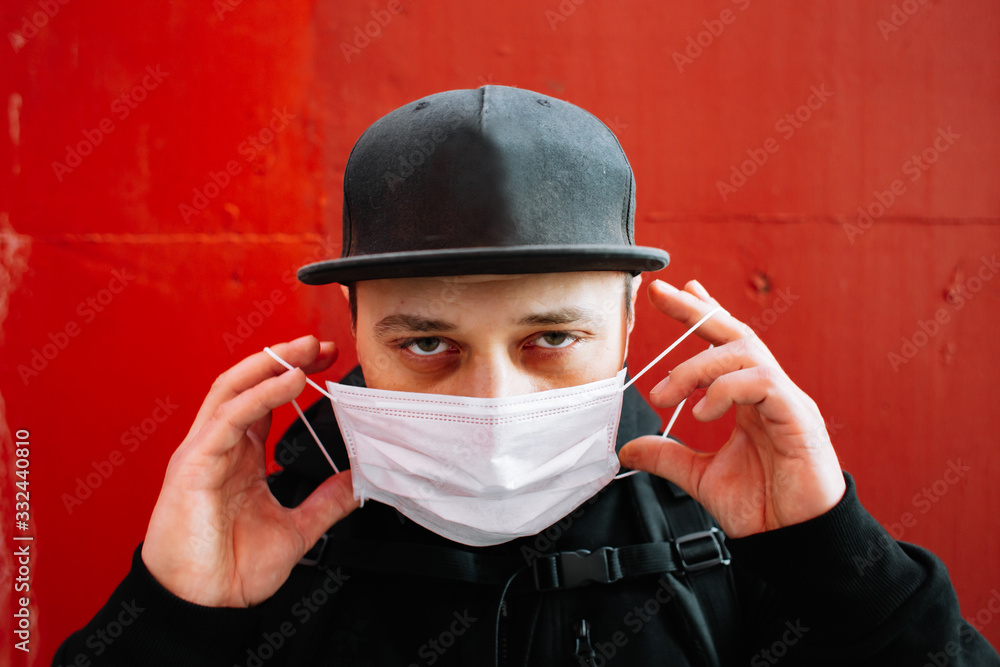 a young man in black sportswear and a baseball cap with a medical mask on his face near the red wall. a young man in black sportswear and a baseball cap with a medical mask on his face near the red wall.