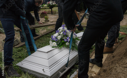 Raasiku / Estonia - January 02 2020: Funeral ceremony in small Lutheran graveyard. Undertakers lift coffin into the grave that is decorated with spruce branches. Flower wreath on wooden white chest. 