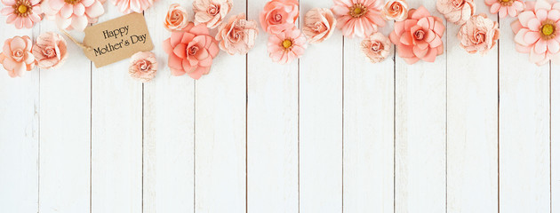 Happy Mothers Day top border with pink paper flowers and gift tag. Above view over a white wood banner background. Copy space.
