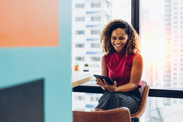 Portrait of cheerful black skin african woman smiling and using tablet for video conversation while relaxing on desk in modern office. Concept of young business people working at home.