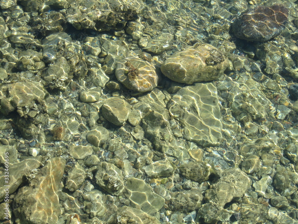 Through the clear water in the rays of the reflection of the sun, you can see the shallow sea floor of sand, pebbles and stones of a gray brown color. Through the clear water in the rays of the reflection of the sun, you can see the shallow sea floor of sand, pebbles and stones of a gray brown color.