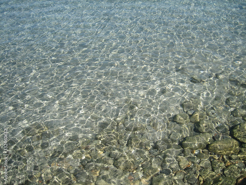 Through the clear water in the rays of the reflection of the sun  you can see the shallow sea floor of sand  pebbles and stones of a gray brown color.