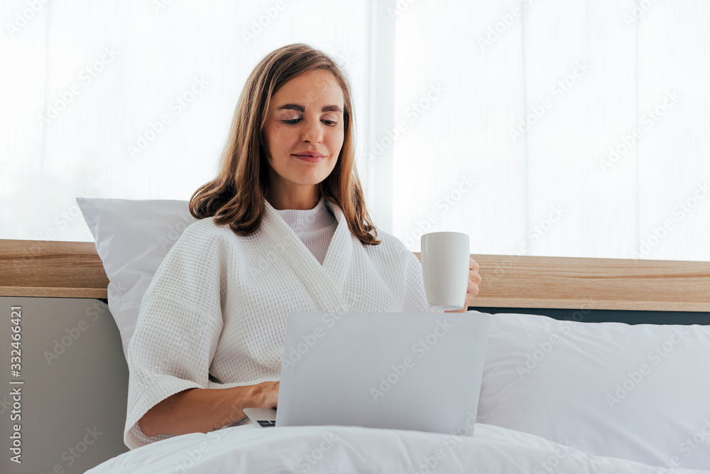 Young joyful woman in pajamas in bedroom holding a cup of coffee - Caucasian girl sits with comfort using computer laptop - Work from home concept Young joyful woman in pajamas in bedroom holding a cup of coffee - Caucasian girl sits with comfort using computer laptop - Work from home concept