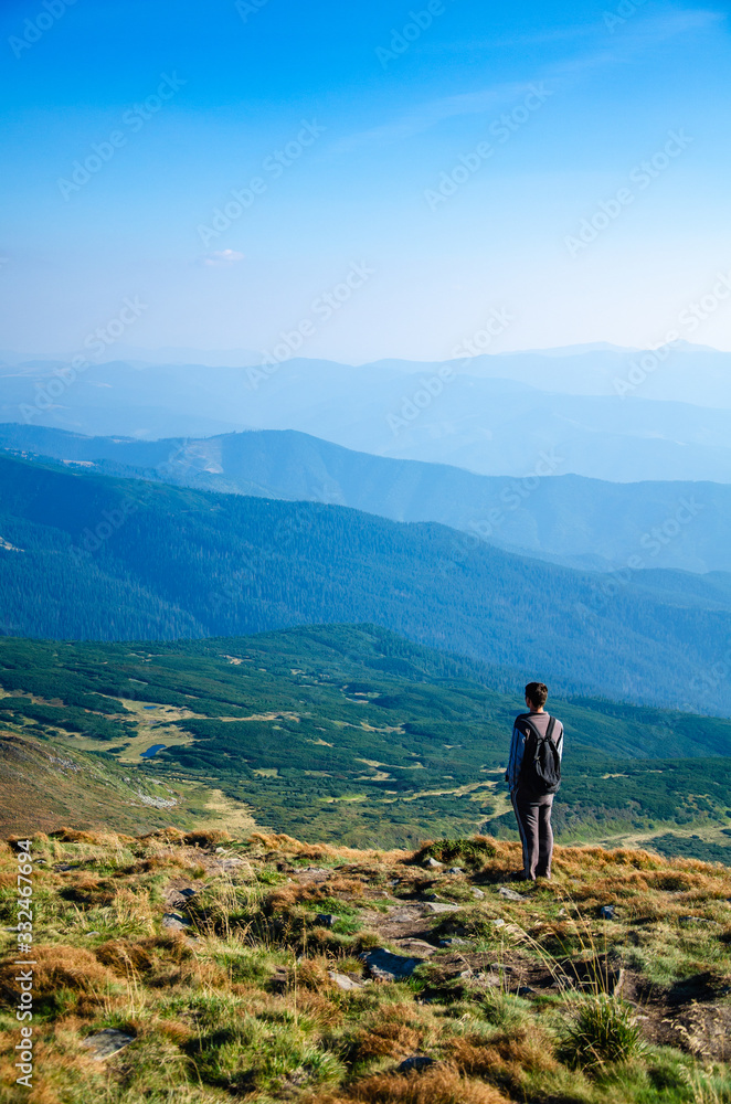 Young man standing on top of a mountain and looks at scenic landscape of ridges and blue sky