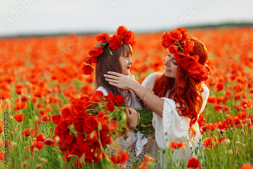 Little girl with redhead mother in white dresses makes wreath on poppy field at summer sunset © onphotoua